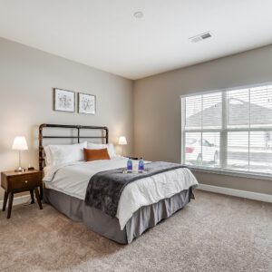 A queen bed in a bedroom with large windows, wall art, and nightstands in a furnished apartment in Westerville Ohio