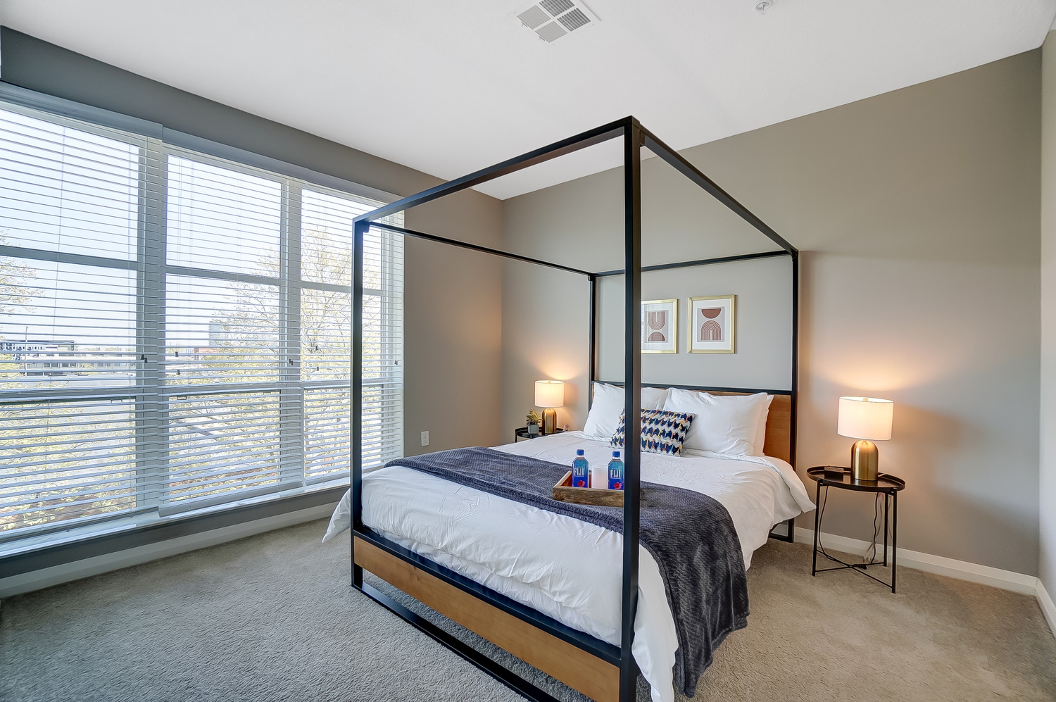 A queen bed in a bedroom with large windows, wall art, and nightstands in a furnished apartment in Downtown Columbus