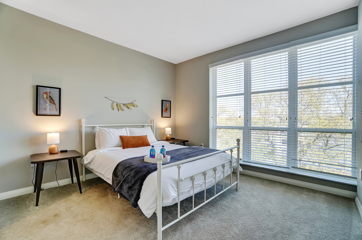 A queen bed in a bedroom with large windows, wall art, and nightstands in a furnished apartment in Downtown Columbus