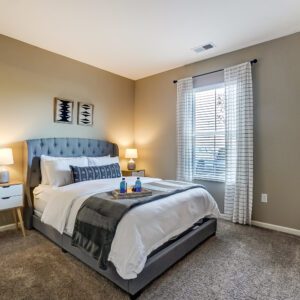 A queen bed in a bedroom with large windows, wall art, and nightstands in a furnished apartment in Hilliard Ohio