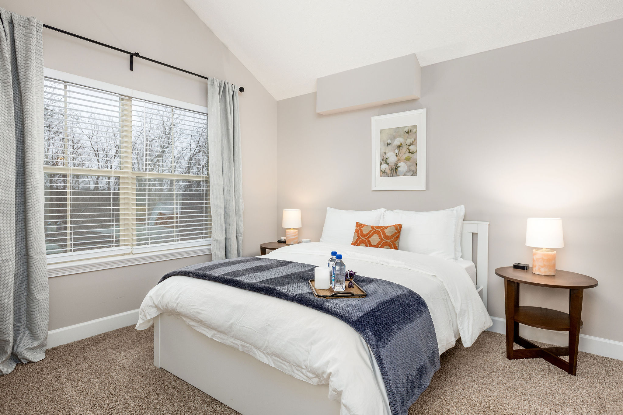 A queen bed in a bedroom with large windows, wall art, and nightstands in a furnished apartment in Westerville Ohio