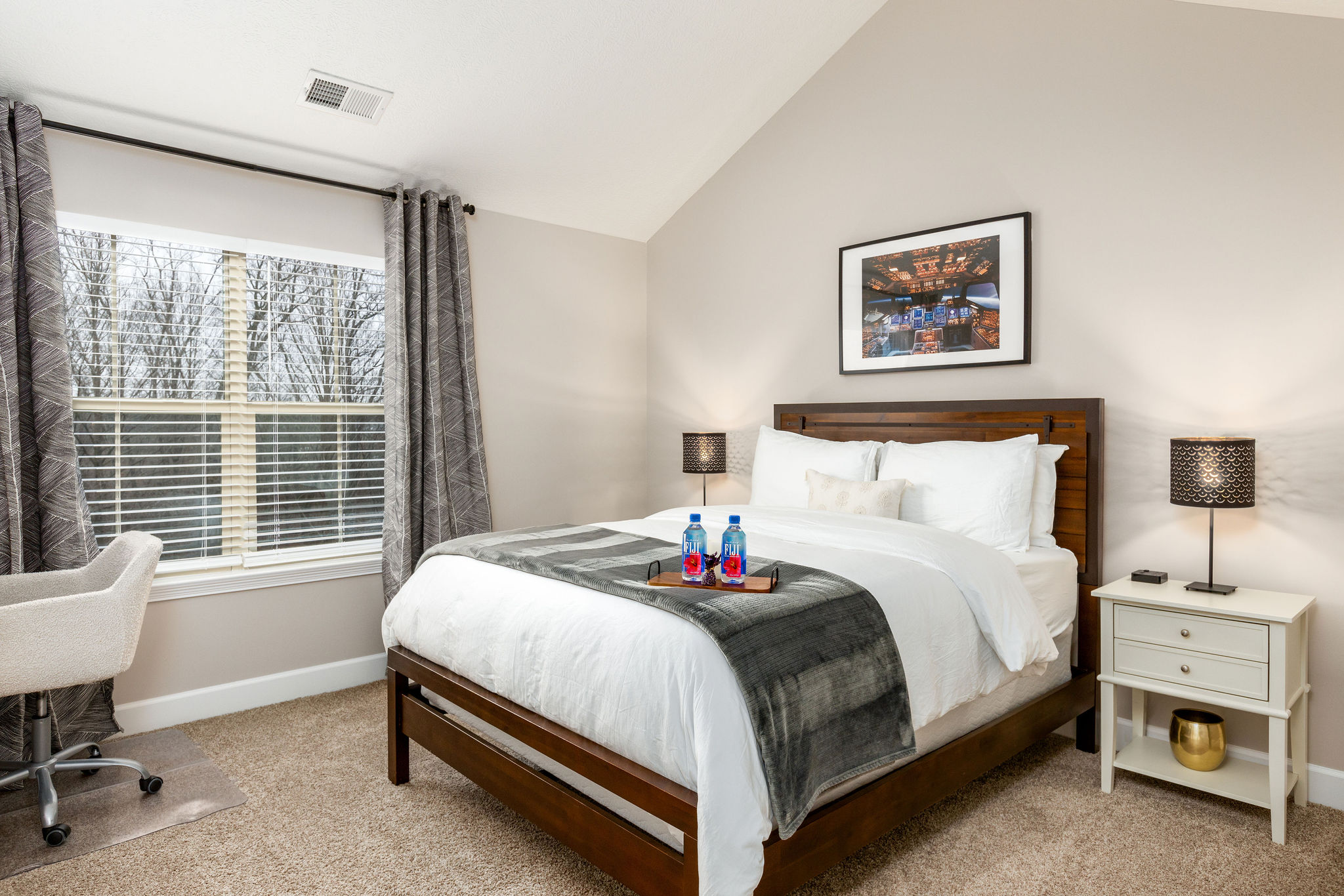 A queen bed in a bedroom with large windows, wall art, and nightstands in a furnished apartment in Westerville Ohio