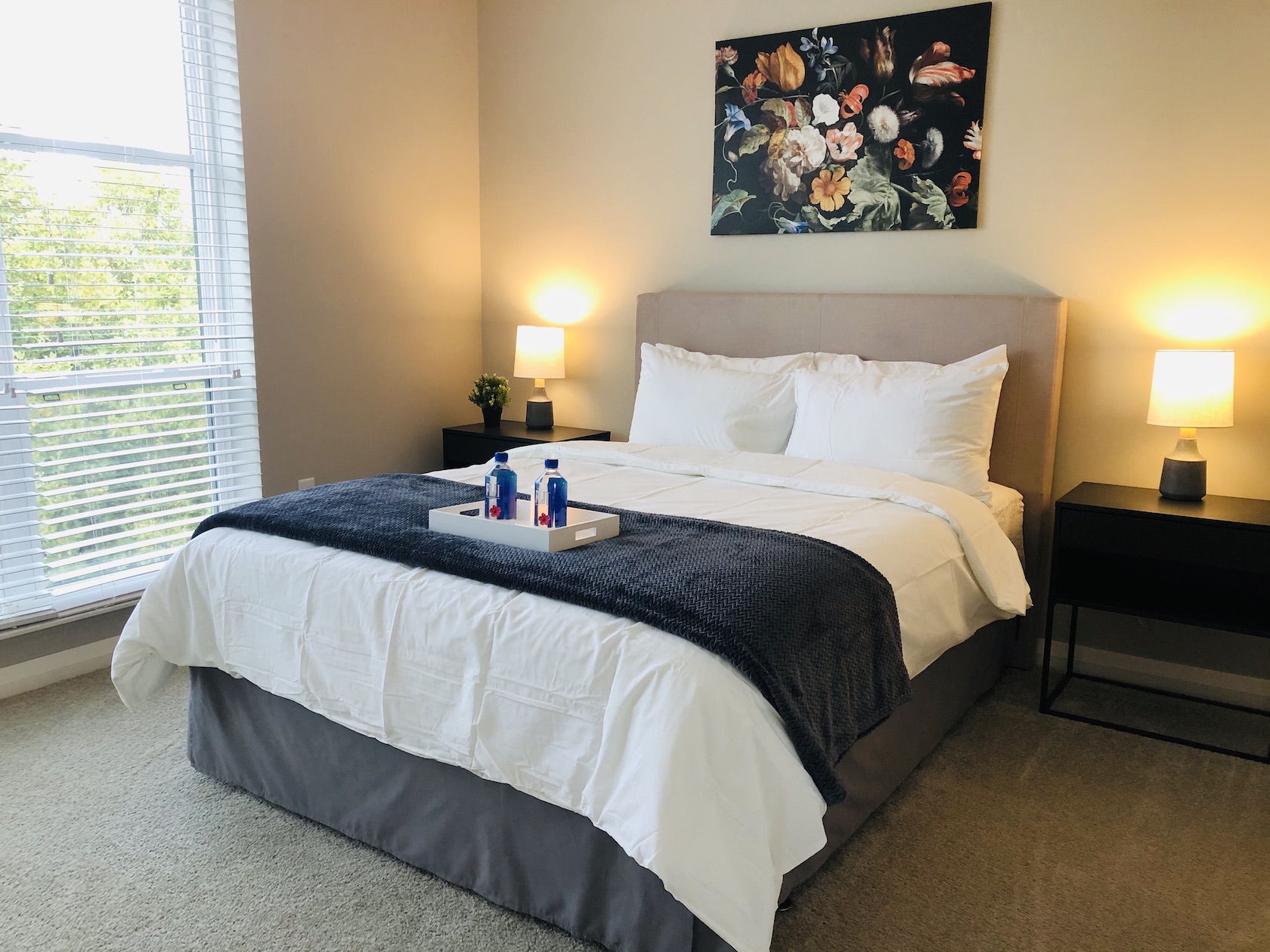 A queen bed in a bedroom with large windows, wall art, and nightstands in a furnished apartment in Downtown Columbus